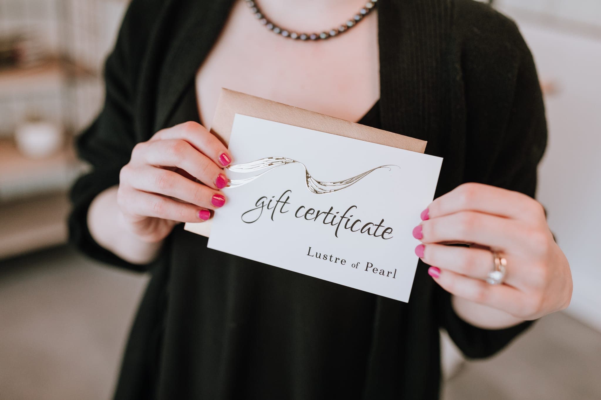 A Lustre of Pearl gift certificate and its envelope, held against a black cardigan and a strand of pearls