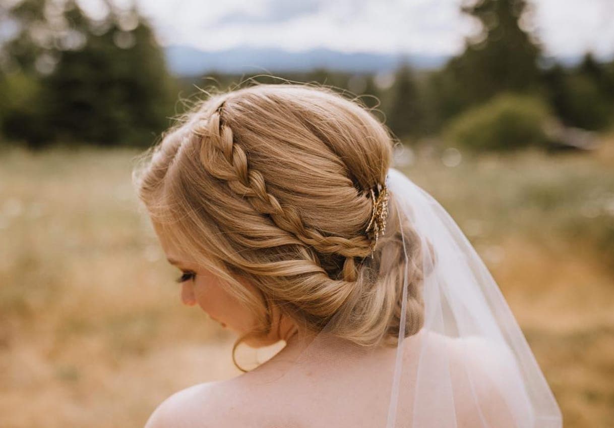 Bride with a side braid sweeping into a low rolled updo beneath a veil, in a golden field