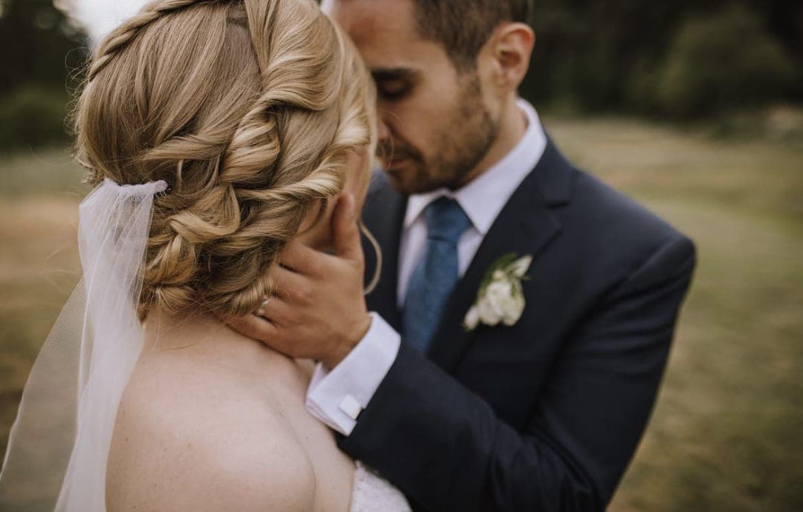 Bride with a braided blonde updo and veil, the groom resting his head against hers
