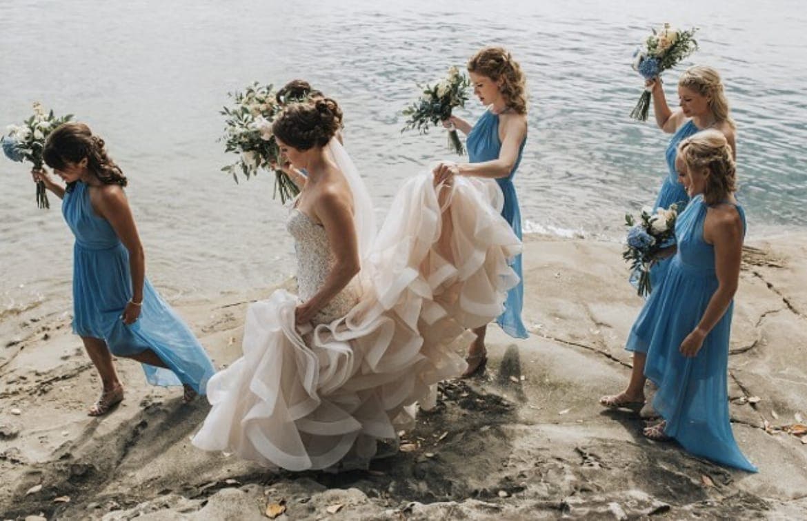 Bride in a ruffled gown and four bridesmaids in blue walking a rocky shoreline with their bouquets