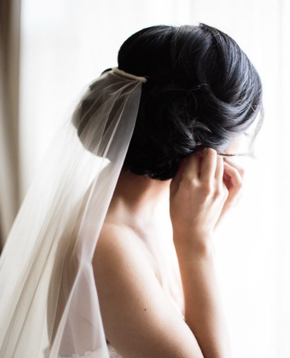 Bride with a full dark chignon and veil, fastening an earring by a window