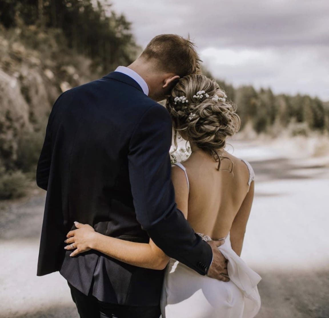 Bride with a low curled updo pinned with small white blossoms, held close by the groom on a coastal road