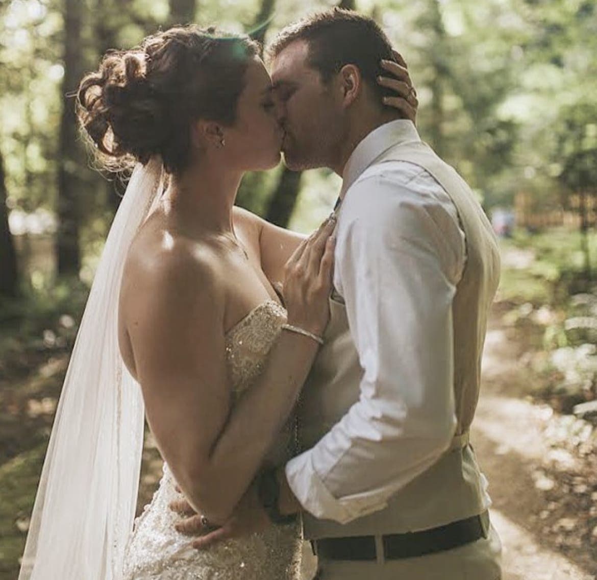 Bride with a pinned-curl updo and long veil kissing the groom on a forest path
