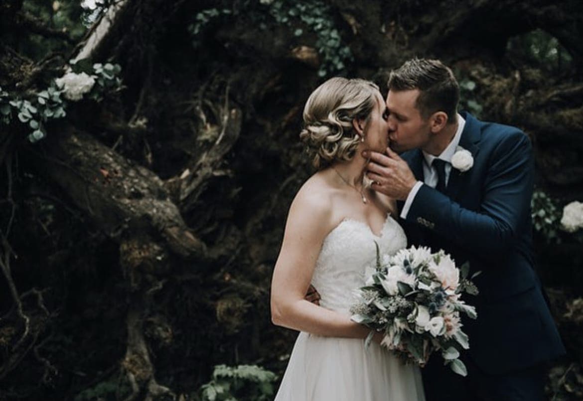 Bride with a blonde updo of sculpted curls kissing the groom, her bouquet in hand, before a dark tangle of tree roots
