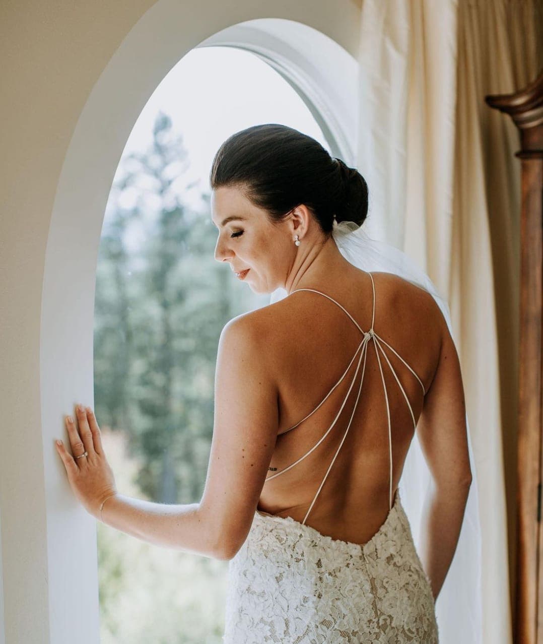 Bride with a sleek dark low bun at an arched window, in a backless lace gown