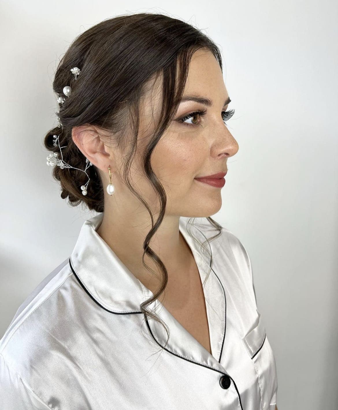 Profile of a bride in a satin robe, her dark low updo set with pearl pins and soft curled tendrils