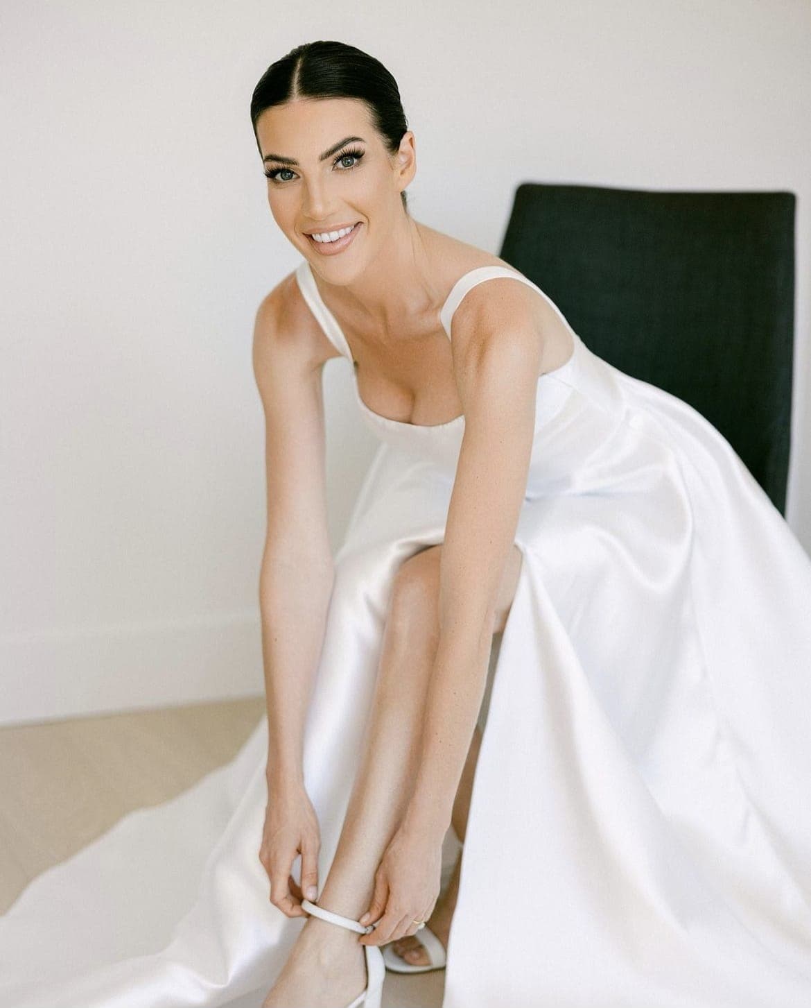Bride in a satin gown with a sleek centre-parted low bun, fastening her shoe
