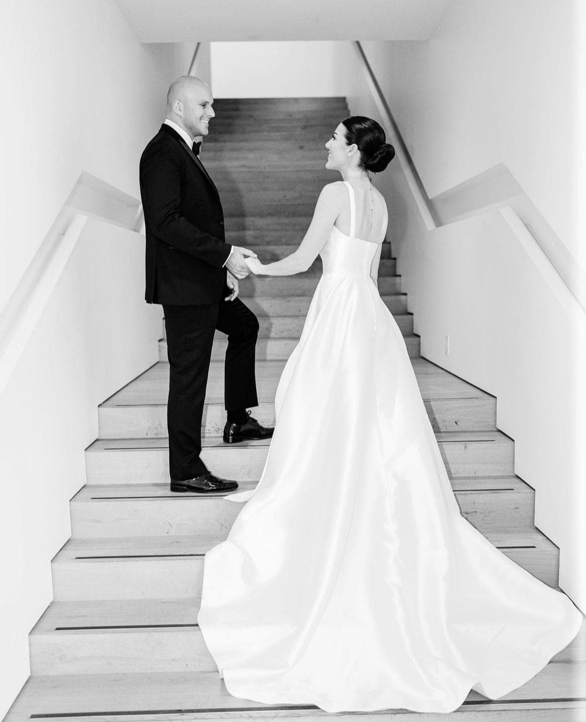 Black-and-white photo of a bride with a low chignon taking the groom's hands on a staircase