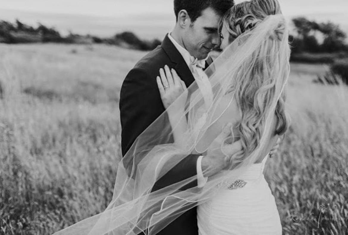 Black-and-white photo of a bride with long blonde curls, forehead to forehead with the groom in a field of tall grass, her veil blown across them