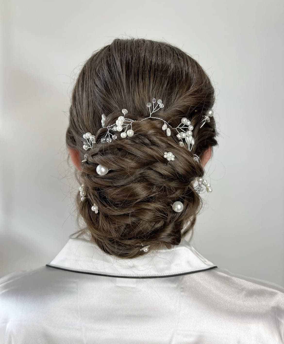 A dark low updo of woven twists from directly behind, dotted with pearl pins beneath a pearl hair vine, above the collar of a satin robe